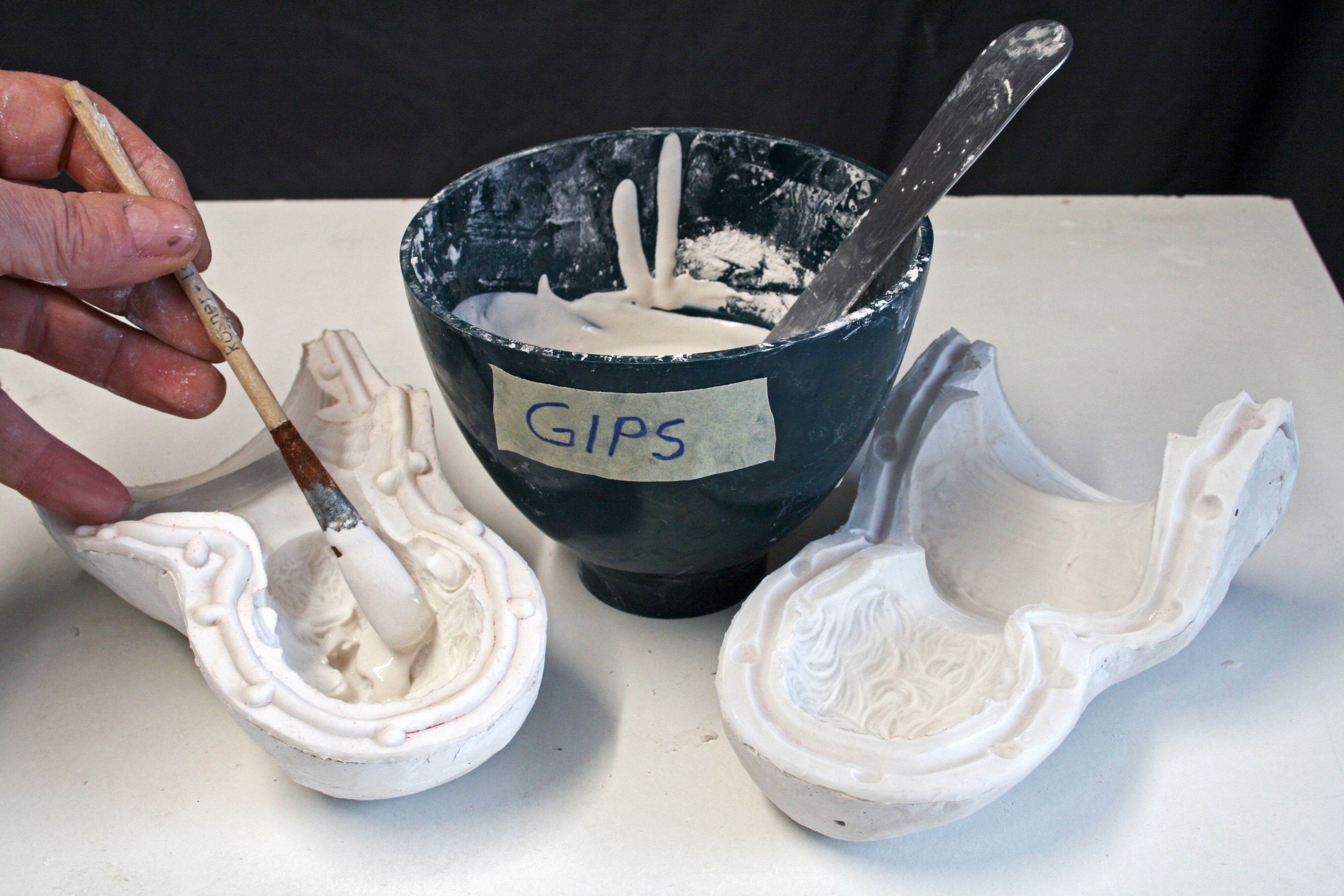 Eine Gipsschale, daneben zwei Formteile. Eine Hand pinselt einen Teil aus. 